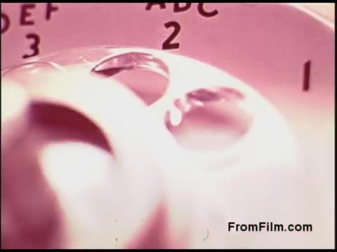 The still from the 1970s Bell System film depicts a close-up view of a rotary dial telephone. The dial is marked with numbers and letters, showcasing the retro design characteristic of that era. The focus on the dial suggests an emphasis on communication technology, highlighting its significance in the context of telephony during that time. The overall color scheme is soft, adding to the vintage feel of the image.