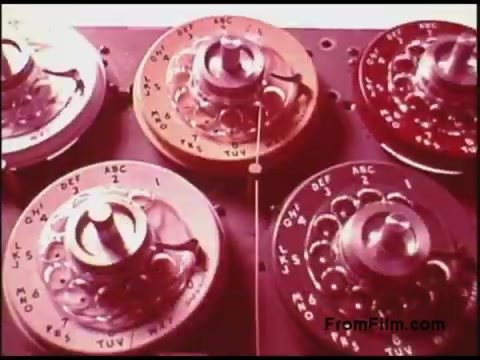 The still from the 16mm 'Bell System Film from the 1970s' features a close-up of a series of rotary switches or dials. The dials are labeled with various letters and numbers, suggesting they are used for technical operations, possibly related to telecommunications or broadcasting. The color palette includes shades of red and pink, giving it a vintage aesthetic, characteristic of films from that era. The focus on the machinery emphasizes the technological context of the film.