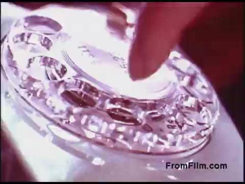 The still features a close-up of a vintage rotary telephone from the 1970s. A finger is poised over the dial, ready to rotate it, showcasing the classic design with circular finger holes and a shiny surface. The lighting gives the scene a nostalgic glow, reflecting the era's technological aesthetics.
