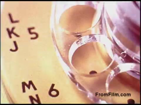 The still features a close-up view of a circular dial or component with engraved letters and numbers, suggesting it is part of a telecommunication device or mechanism. The materials appear metallic and glass-like, reflecting the design aesthetics typical of technology from the 1970s. The overall composition highlights the intricate details of the machinery, indicative of the era's engineering.