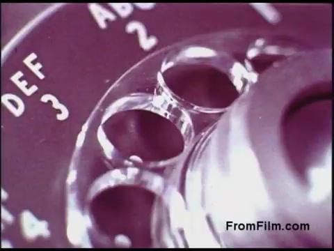 The still depicts a close-up view of a rotary dial from a vintage telephone, focusing on the numbers and letters engraved on the dial. The image is likely vibrant and colorful, characteristic of film from the 1970s, emphasizing the design and mechanics of telecommunication technology from that era. The retro aesthetic is evident, highlighting the nostalgia associated with older communication methods.