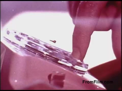 The still depicts a close-up shot of a person's finger poised above a transparent rotary telephone dial, typical of the 1970s. The composition emphasizes the dial's circular shape and grooves, suggesting a moment of interaction or dialing. The colors are slightly muted, reminiscent of film stock from that era. The background is minimal, focusing attention on the hand and the phone component, evoking a sense of nostalgia for the technology of the time.