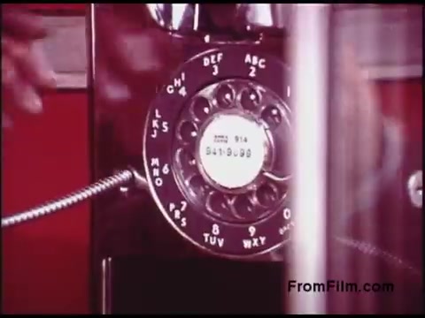The still features a vintage rotary telephone prominently displayed. It has a glossy finish with a distinct circular dial, numbered 0-9, including letters associated with each digit. A hand can be seen reaching for the dial, suggesting an action of placing a call. The background is a muted red, characteristic of the era's design aesthetic. This film likely captures a slice of life from the 1970s, emphasizing the telephone's role in communication during that time.