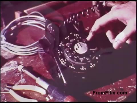 The still shows a close-up of a rotary dial telephone on a surface, likely a workbench. A hand is poised over the dial, preparing to turn it, while a coiled phone cord and a couple of tools—pliers and a screwdriver—are visible in the foreground. The scene likely depicts a repair or installation process related to telecommunications, characteristic of Bell System operations from the 1970s. The colors are vivid, reflecting the film's age and providing a nostalgic feel.