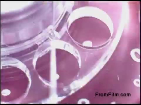 The still features close-up imagery of a clear film reel, likely from a 16mm film, set against a blurred, colorful background. The intricate details of the reel's holes and structure are highlighted, emphasizing the texture and design typical of film equipment from the 1970s. The overall aesthetic reflects the era’s technological advancements in film production.