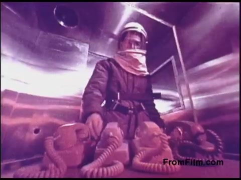 The still from the 1970s Bell System film features a person in a protective suit and helmet, positioned in a metallic environment. The color palette has a distinctive purple tint, adding an otherworldly effect. The individual is surrounded by equipment with coiled cords, suggesting a connection to communication or technology. The setting appears to be laboratory-like, emphasizing a focus on safety and advanced technology.