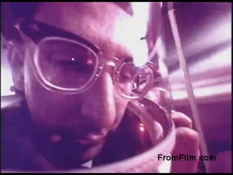 The still captures a close-up of a man wearing glasses and an industrial helmet, possibly in a space or scientific setting, highlighting a moment of concentration or reflection. The image is likely characterized by the distinct color grading typical of 1970s film, with a slightly muted or vintage look, emphasizing its period authenticity. The context suggests a focus on technology or experimentation related to Bell System's projects.