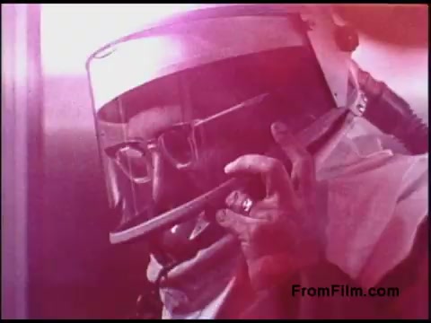 The still from the 'Bell System Film from the 1970s' features a man wearing a protective helmet and glasses, holding a telephone receiver. The image has a soft, reddish tint, suggesting a dramatic or stylized presentation. The setting appears to involve a technical or industrial environment, reflecting the era's focus on safety and communication technology.