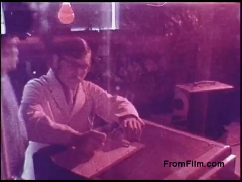 The still depicts a man in a white laboratory coat, engaged in a task at a workbench. He appears focused on a small object, surrounded by various items, including a vintage box and hanging light bulbs. The overall scene has a pinkish hue, suggesting lighting or color grading typical of the 1970s. The background hints at a lab or workshop environment, emphasizing a scientific or experimental context.