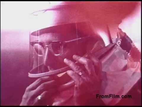 The still from the 1970s Bell System film features a man in a protective suit, complete with a helmet. He appears to be communicating or inspecting something through a visor. The color tones are warm, with a reddish hue that adds a vintage feel to the image. The subject's expression conveys focus and concentration, suggesting a serious or technical context in the film.