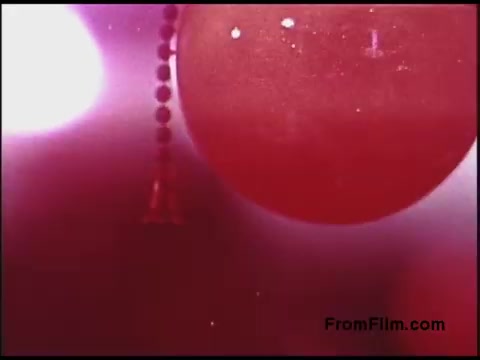 The still depicts a close-up of a red background featuring a circular shape that could be a balloon or similar object. There are beads or a string-like element hanging, contrasting with the soft focus of the surrounding pink and white light. The overall aesthetic suggests a dreamy, abstract quality typical of 1970s experimental film.