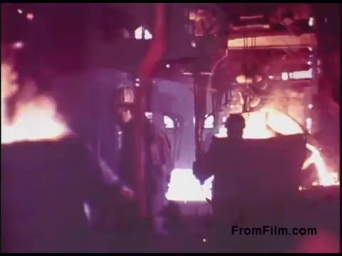 The still from the 'Bell System Film from the 1970s' depicts a factory or industrial setting, characterized by the presence of intense flames and glowing metal. Workers are engaged in manual tasks amidst a backdrop of machinery and equipment, illuminated by warm, harsh lighting. The overall atmosphere conveys a sense of labor and industrial activity. The color palette suggests a vintage aesthetic, typical of film from that era.