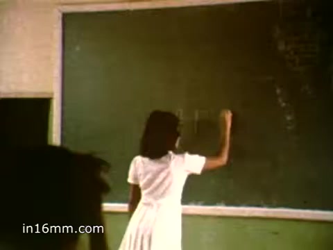 The still from the 1980s PSA/advert for Plan International USA shows a young girl in a white dress writing on a green chalkboard. The setting appears to be a classroom, emphasizing education as a vital component in the fight against poverty. This aligns with Plan International's mission to improve the lives of children globally by providing opportunities for education and support.
