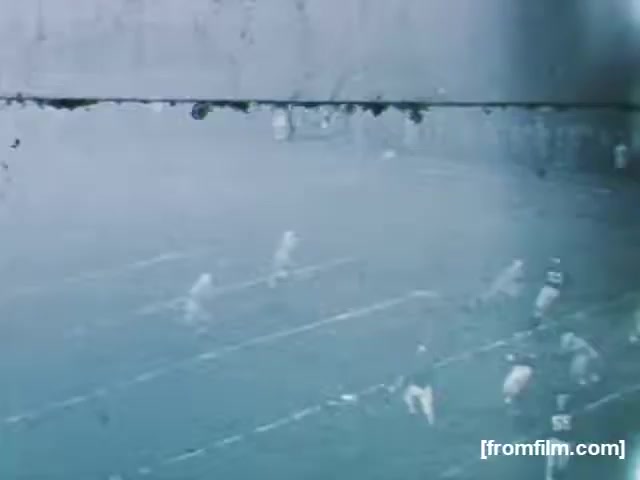 The still shows a high school football game from 1961, featuring players on a foggy field. The image captures the moment of play, with athletes in action and a blurred background, suggesting a lively sporting event. The scene may reflect the atmosphere of high school sports during that era.