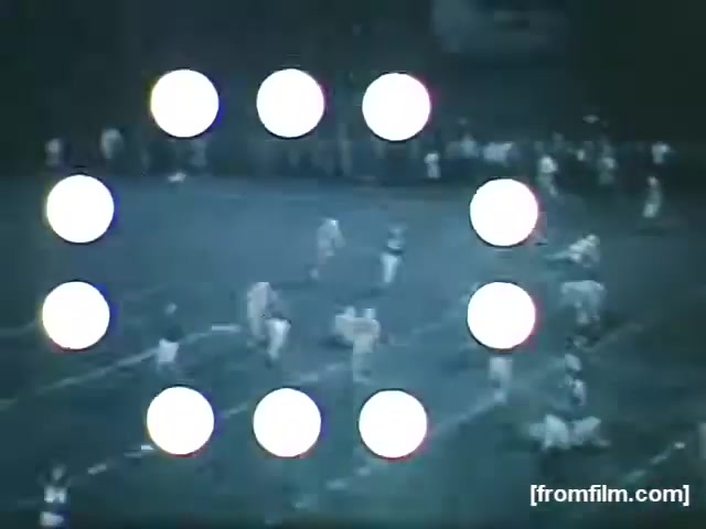 The still features a night scene from a high school football game in 1961, with players on the field engaged in action. The image is overlaid with a graphic display of circular markers, possibly for framing or effect. The location and teams are unknown, but it captures the energy and atmosphere typical of high school football, highlighting the sport's community spirit.