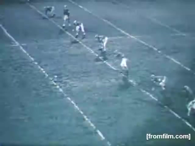 The still from 'High School Football - 1961 (2)' shows a distant view of a football field during a game. Players from two teams are positioned for a play, with some players in a huddle and others lined up along the line of scrimmage. The field appears muddy, indicative of the weather or conditions during the game. The image is in a vintage style, reflecting the era of 1961.