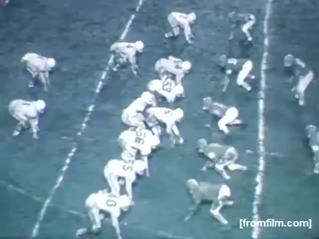 The still from 'High School Football - 1961 (2)' shows a football game featuring two teams in action. The players are positioned in a formation on the field, with one team on the offensive and the other on the defensive. The scene captures the classic atmosphere of high school football, complete with players in traditional football gear and a distinct, gritty playing field. The image is in black and white, emphasizing the era's vintage quality.