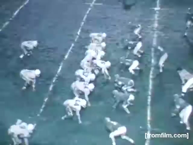 The still shows a high school football game from 1961, featuring players lined up on a muddy field. The teams are in contrasting colored uniforms, with players in a ready stance on both sides of the field, preparing for the snap. The perspective highlights the alignment and anticipation typical of a football play.