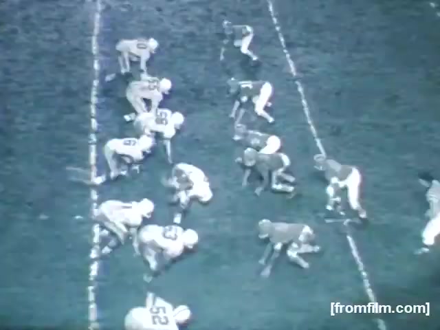 The still shows a high school football game from 1961, featuring players lined up on the field. The scene captures a moment just before the snap, with both teams positioned in a typical offensive and defensive formation. The players are wearing classic football uniforms, typical of the era, with contrasting colors. The grass field appears well-worn, indicating the intensity of the game.
