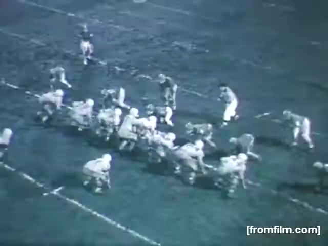 The still from 'High School Football - 1961 (2) [FF0005]' depicts a high school football game in black and white. The players are in an active formation on the field, with half the team crouched in front and the other half positioned behind a line of scrimmage. The setting suggests an early evening game, with visible yard lines and a textured grass field. The overall composition captures a moment of tension and anticipation typical of football matches.