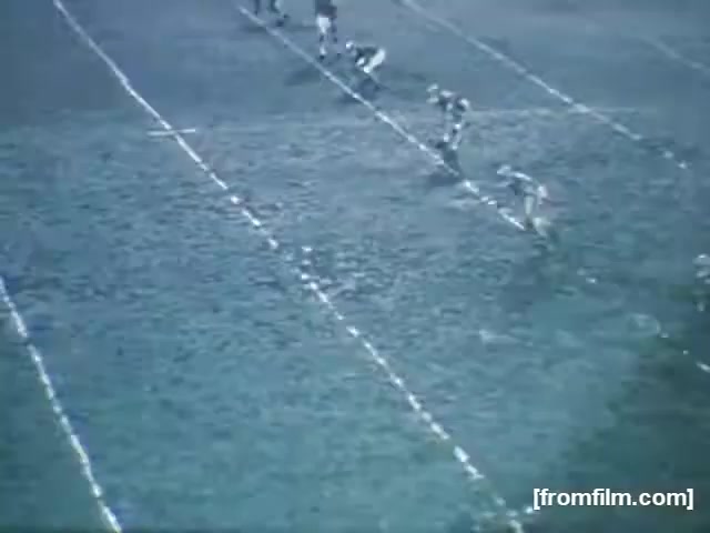 The still depicts a high school football game from 1961. Players are lined up on the field, which shows the markings typical of a football field. The image features a slightly faded quality, characteristic of 16mm film. The scene captures the pre-play moment, emphasizing the players’ readiness for action in an unknown location and with unidentified teams.