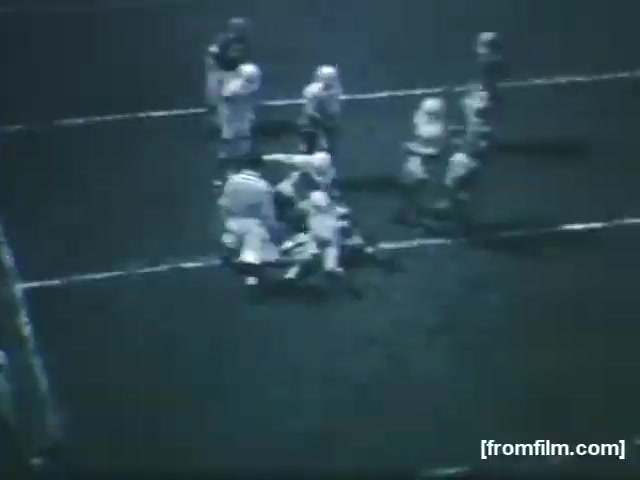 The still captures a moment from a high school football game in 1961, showing players in action on the field. The scene is monochromatic, typical of 16mm film of the era, and features a group of players clustered around a central play, possibly involving a tackle. The atmosphere conveys the intensity and camaraderie of high school sports.