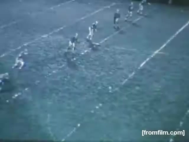 The still captures a high school football game from 1961, showcasing players on a muddy field. The perspective suggests a play about to unfold, with several players positioned strategically for action. The scene conveys the competitive spirit of youth sports, emphasizing teamwork and athleticism amid challenging conditions.