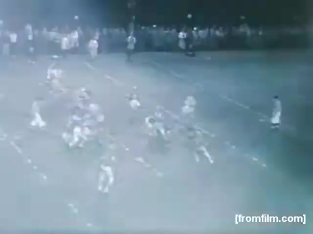 The still from 'High School Football - 1961 (2)' captures a nighttime football scene. Players in various uniforms are engaged on the field, with some in motion. A large crowd is visible in the background, indicating a lively atmosphere. The lighting gives a slightly hazy effect, typical of older film stock. The specific location and teams are unknown.