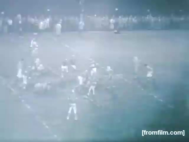 The still from 'High School Football - 1961 (2)' captures a nighttime football game. Players in contrasting uniforms are engaged in a play on a well-marked field, surrounded by an audience in the background. The lighting is dim, contributing to a vintage atmosphere typical of the era.