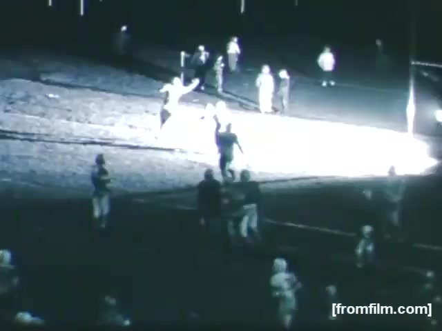 The still captures a nighttime high school football scene from 1961. Players are on the field, with some celebrating a play. The illumination comes from a nearby light source, casting bright lighting over part of the field while others remain in shadow. The atmosphere suggests excitement and energy typical of a high school football game.
