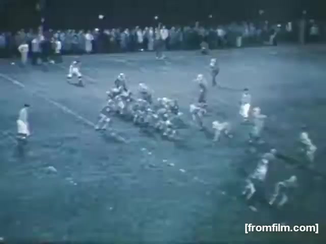 The still shows a scene from a high school football game in 1961, featuring players on the field in a night setting. The focus is on a group of players positioned in a formation, preparing for a play. In the background, a crowd of spectators watches the game. The lighting suggests a night game atmosphere, highlighting the players in action.