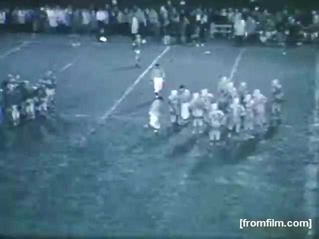 The still captures a high school football game from 1961, featuring two teams on the field. The players are gathered in a huddle, discussing their next play, while spectators watch from the sidelines in the background. The field appears muddy, indicating challenging playing conditions. The scene conveys a sense of competition and camaraderie typical of high school sports.