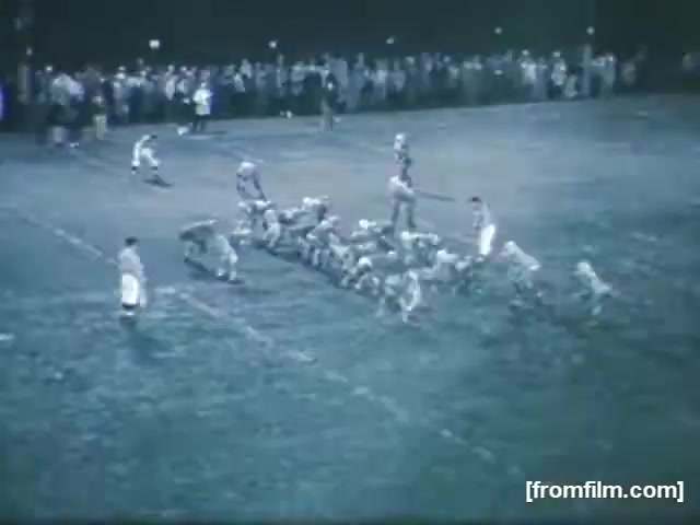 The still from 'High School Football - 1961 (2)' captures an evening football game on a grassy field. A team is in formation on the left, preparing for a play, while a few players stand ready to snap the ball. In the background, a crowd is gathered, illuminated by lights, creating a lively atmosphere typical of high school sports. The overall scene reflects the excitement and community engagement of local football matches during that era.