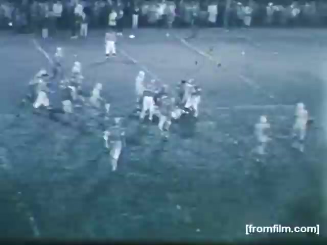 The still shows a high school football game set in an evening atmosphere. Players are engaged in a dynamic play on the field, with one quarterback poised to throw the ball while teammates and opponents are in motion. Spectators can be seen in the background, indicating a lively game atmosphere. The black-and-white visual adds a nostalgic tone, characteristic of footage from 1961.