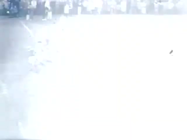 The still from 'High School Football - 1961 (2)' appears to depict a scene from a football game, likely in a high school setting. The image is quite bright, suggesting overexposure, which obscures details. In the foreground, there are players engaged in action, while a crowd can be seen in the background. The overall context indicates an active sports event, emphasizing the excitement of high school football in the early 1960s.