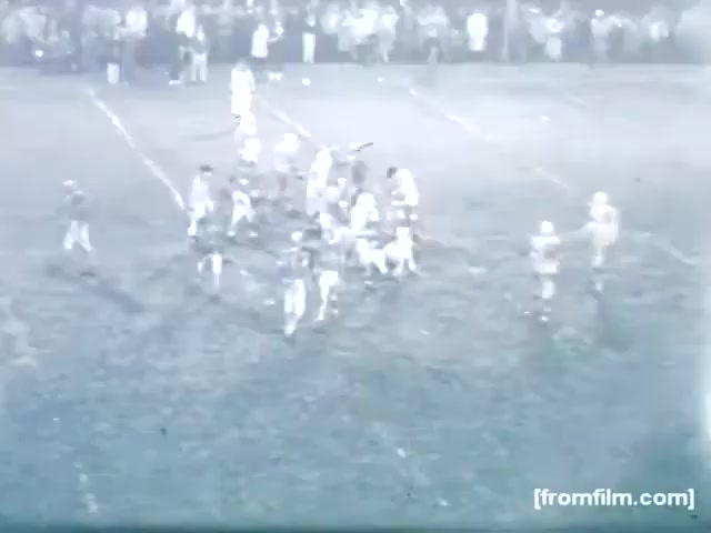 The still captures a moment from a high school football game in 1961. The scene depicts a group of players on the field, with one team huddled in a play formation. The field shows signs of wear, and spectators can be seen in the background, indicating a lively atmosphere. The image has a vintage feel characteristic of 16mm film.