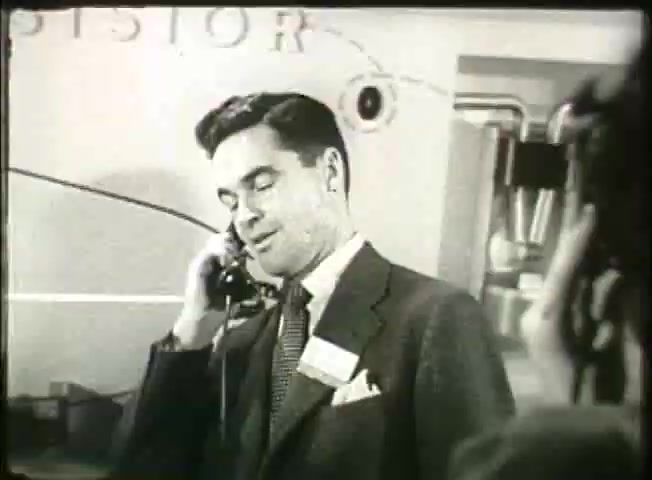The still from 'Story Without End' features a man in a suit speaking on the phone, likely in a central office setting. He appears engaged and professional, reflective of the communication technology and business environment of the late 1940s. The background suggests a technical or informational context, possibly illustrating advancements in the Bell System network at that time. The film aims to showcase the evolution of telecommunications and the significance of such technology in everyday life.