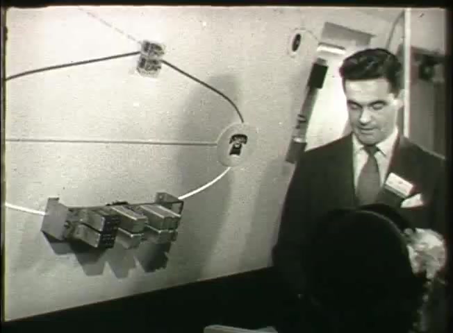 The still from the Bell System film 'Story Without End' depicts a man in a suit standing before a wall-mounted demonstration of telecommunications equipment. The display features a series of connected devices, possibly switches or relays, illustrating the inner workings of a telephone network. The setting appears to be a central office, emphasizing the technical aspects of communication systems in the late 1940s. The man appears to be explaining the setup, likely representing advancements in telecommunications during that era.