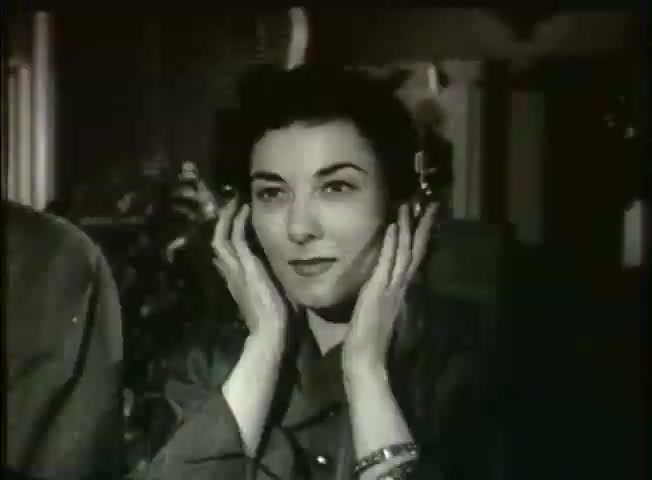 The still from 'Story Without End,' a Bell System film from the late 1940s, depicts a woman wearing a headset, attentively listening. She has a thoughtful expression, with her hands gently placed on her ears, suggesting engagement with what she hears. The surroundings imply a central office setting, reinforcing the film's focus on telecommunications and the functionality of the network during that era. The vintage aesthetic highlights the technological context of the time.