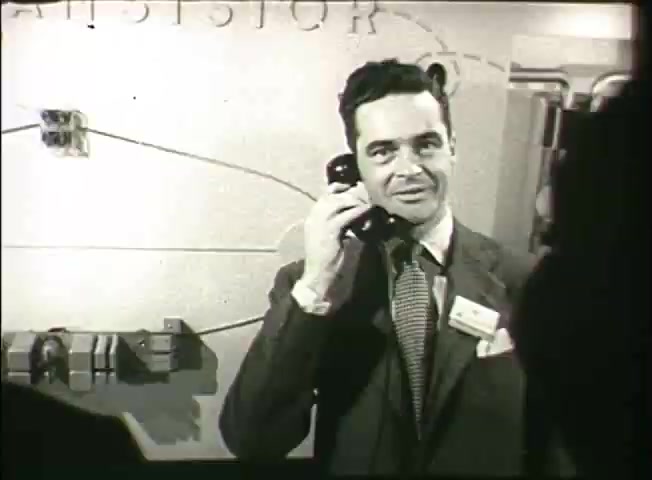 The still from 'Story Without End' features a well-dressed man speaking on a vintage telephone. He appears to be engaged in a conversation while smiling, conveying a sense of professionalism and enthusiasm. The background includes a wall with wiring and diagrams, likely illustrative of telecommunications concepts from the late 1940s, hinting at advancements in the Bell System network. The overall atmosphere suggests a focus on innovation and the importance of communication technology during that era.