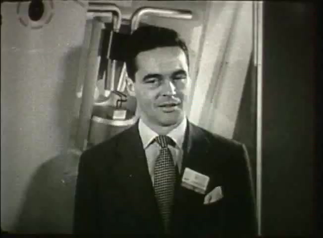 The still features a well-dressed man in a suit and tie, standing in front of some technical equipment, likely related to telecommunications. His expression is friendly and professional, suggesting he may be presenting information. The setting appears to be a central office environment, indicative of the late 1940s Bell System context. The film likely emphasizes advancements in communication technology during that era.
