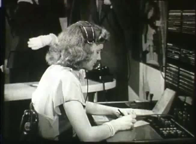 The still from 'Story Without End' shows a female telephone operator in the late 1940s, wearing a headset and sitting at a desk equipped with various communication devices. She is focused on writing notes as a man gestures in the background, indicating an interaction. The setting reflects the era's telecommunication practices and the role of operators in managing calls, providing insight into the Bell System's operations during that time.