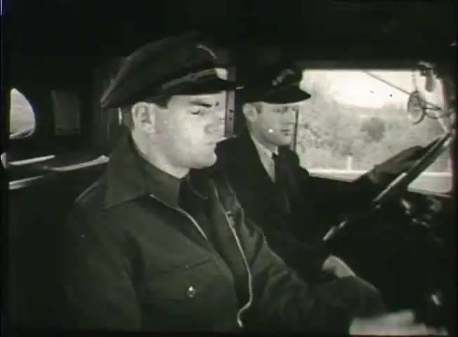 The still from 'Story Without End,' a Bell System film from the late 1940s, features two men in a vehicle's front seat. The driver, wearing a uniform with a cap, appears focused on the road, while the passenger, also in a cap, looks toward him. The setting suggests a context related to telecommunications, likely highlighting the operational aspects of the company during that era. The film captures a moment of teamwork within the Bell System's network infrastructure.