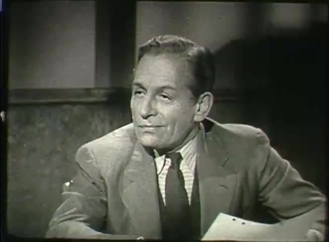 The still from 'Story Without End,' a Bell System film from the late 1940s, features a man in a suit seated at a table. He appears engaged in conversation or reflection, possibly discussing topics related to telecommunications or network operations. The setting suggests a formal environment, likely a central office or meeting room. His expression conveys attentiveness, indicating a serious or informative discussion relevant to the film’s themes.