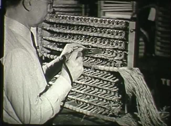 The still from the film 'Story Without End' depicts a technician working on a telephone switchboard, highlighted by intricate wiring. The man, dressed in a formal shirt and tie, is using tools to manipulate wires, showcasing the hands-on nature of telecommunications work in the late 1940s. The background suggests a central office environment, emphasizing the complexity and importance of the network infrastructure at that time. This captures a moment of the evolving technology within the Bell System.