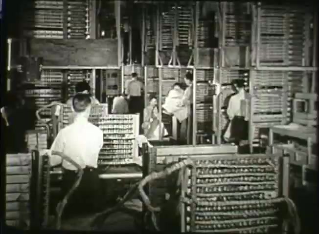 The still from 'Story Without End' features a bustling central office environment, showcasing several workers interacting with large switchboard equipment typical of the late 1940s. The layout includes rows of intricate wiring and machinery, indicating the complexity of the telecommunications infrastructure at the time. Employees, dressed in period-appropriate attire, are seen engaged in various tasks, emphasizing the human element behind the technology. This visual captures the operational side of the Bell System during an era of significant advancement in communication networks.