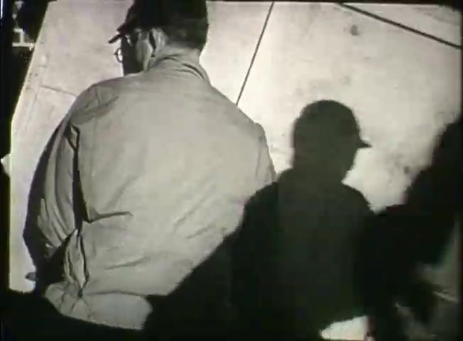 The still from 'Story Without End' shows a worker, possibly in a central office environment, viewed from the back. He wears glasses and a cap, engaged in his task, with shadows cast across the surface. The monochrome presentation emphasizes the era's industrial setting, reflecting the film's focus on the Bell System's operations during the late 1940s, highlighting both personnel and the technical aspects of communication networks.