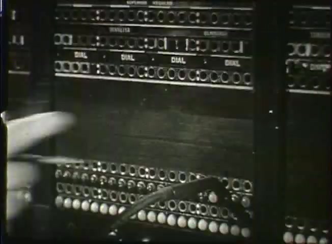 The still features a close-up of an old telephone exchange panel, showcasing a series of labeled dials and rows of holes used for connecting calls. A hand is seen pointing towards the apparatus, emphasizing the manual operation involved in the telecommunications process of the late 1940s. This reflects the technological landscape of the time, before the advent of modern automated systems, highlighting the intricate work within a central office.