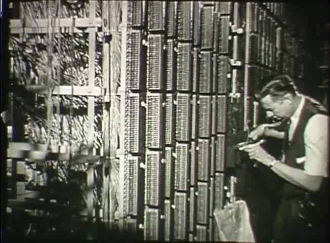 The still shows a technician in a central office environment from the late 1940s, working on a complex array of telephone switching equipment. The backdrop features rows of interconnected panels and wiring, indicative of the Bell System's telecommunications infrastructure at the time. The technician is focused on his task, showcasing the hands-on nature of maintenance and operations in the era's telephone network. The black-and-white image captures a moment of technological precision and the evolving communication landscape.