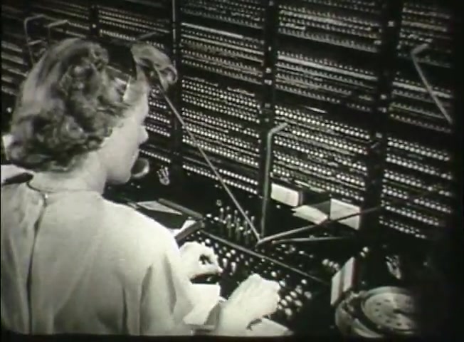 The still from 'Story Without End' depicts a female operator at a telephone switchboard, characteristic of the late 1940s. She is engaged in managing calls, with multiple circuits visible behind her. The scene emphasizes the manual processes involved in telecommunications, reflecting the technology and central office operations of the time. The composition highlights the operator's role in connecting calls, which was crucial before the advent of automated systems.